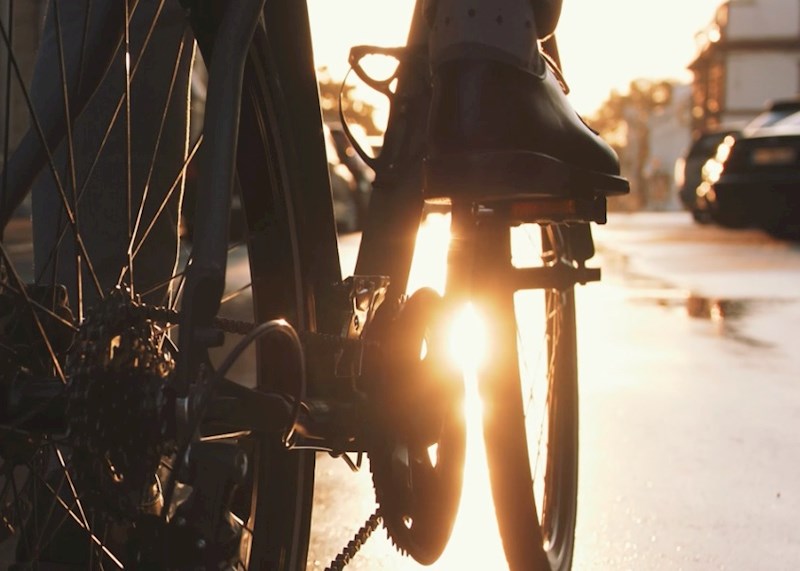 Closeup of a person riding a bicycle that shows the rear tire and the gears, spokes, and chains with the sunset appearing through the pedals.